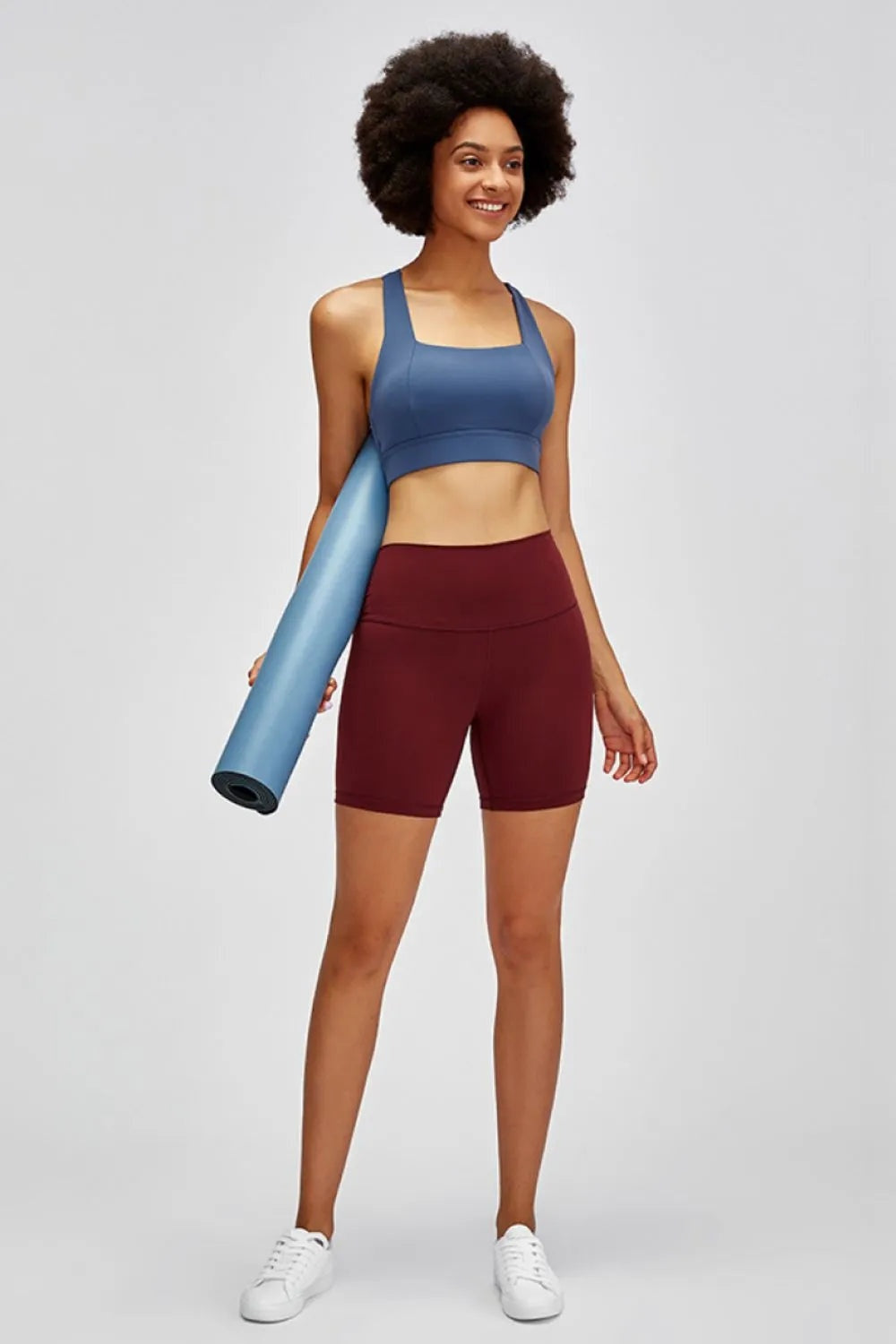 Smiling model wears burgundy high waist training shorts with a wide waistband and fitted, stretchy mid-thigh silhouette while standing and holding a yoga mat.