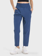 Model wears blue pocketed high-waist active pants with a gathered waistband, relaxed straight-leg silhouette, and hands tucked into the side pockets.