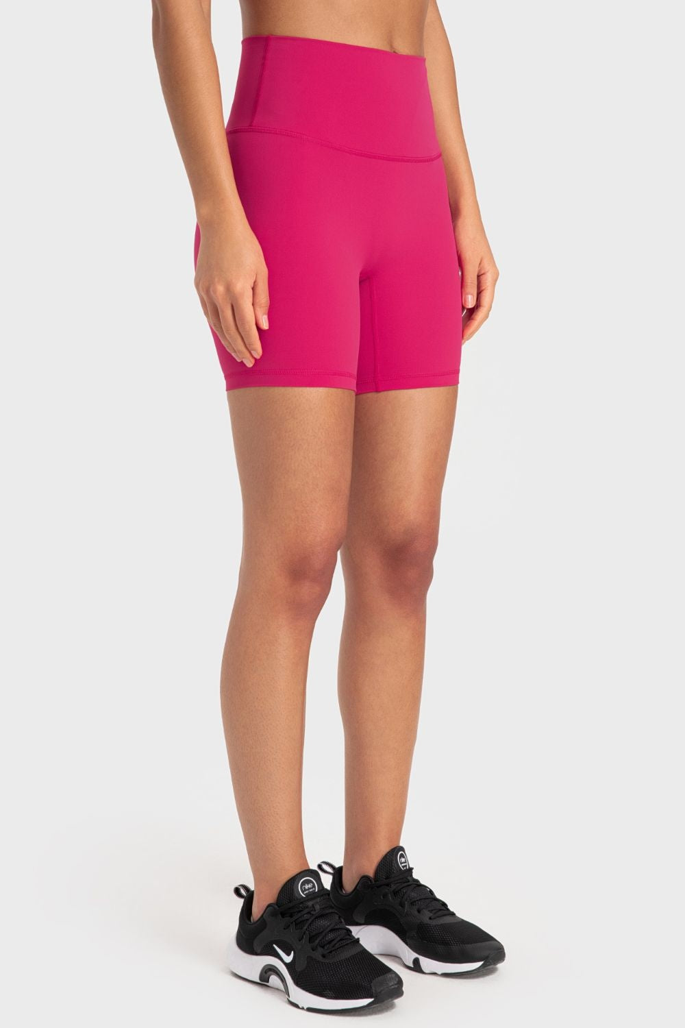 A model stands with arms at her sides wearing solid bright pink biker shorts with a wide waistband, fitted mid-thigh silhouette, and visible front and leg seams.