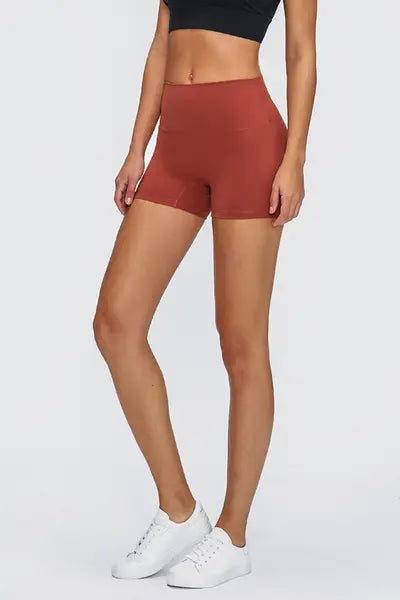 Model wearing rust-red Stitch Active Shorts with a high-rise, stretchy fitted silhouette, smooth waistband, and mid-thigh length, posed standing with one leg bent.
