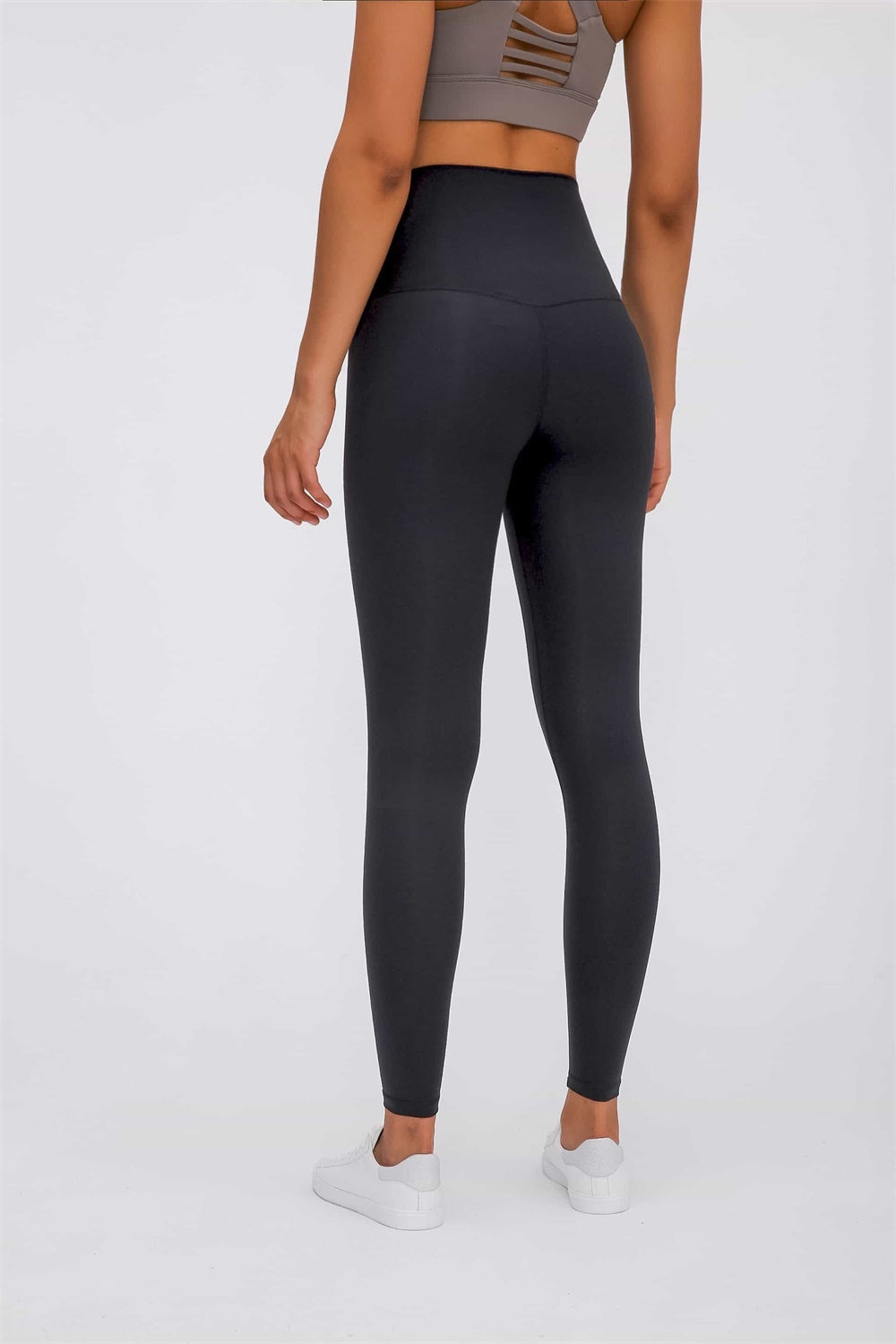 Model shown from behind wearing black high-waist leggings with a wide elastic waistband, smooth construction, and a fitted ankle-length silhouette.