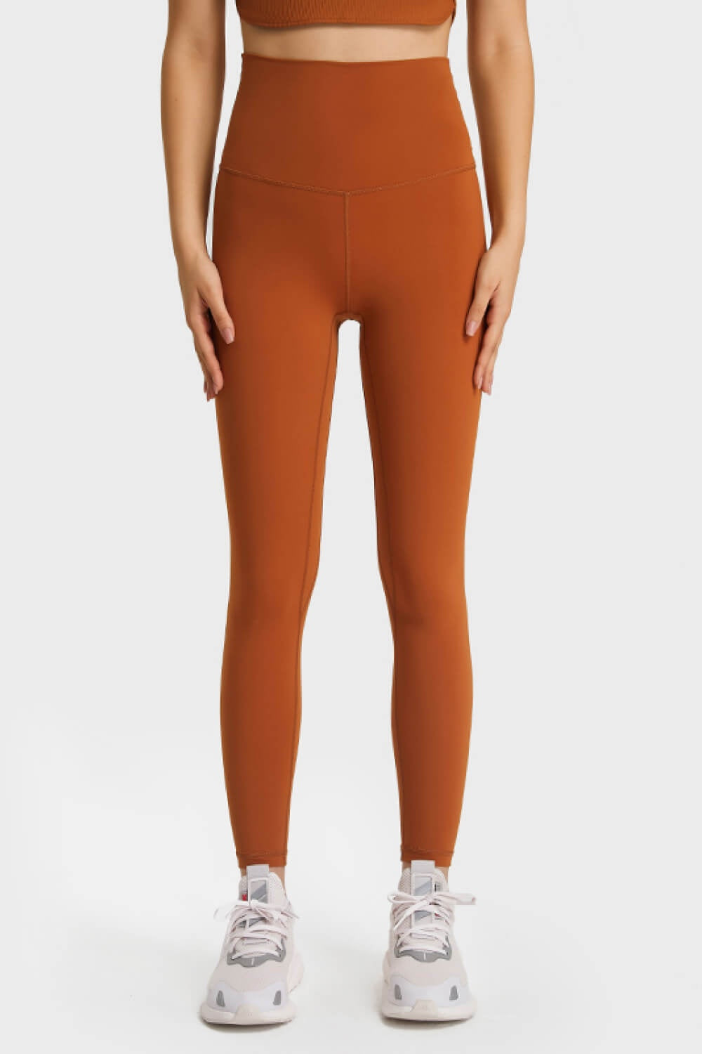 Model stands facing forward in rust-orange high-waist leggings with a smooth, stretchy, slim-fitting silhouette, wide elastic waistband, and visible front seams.