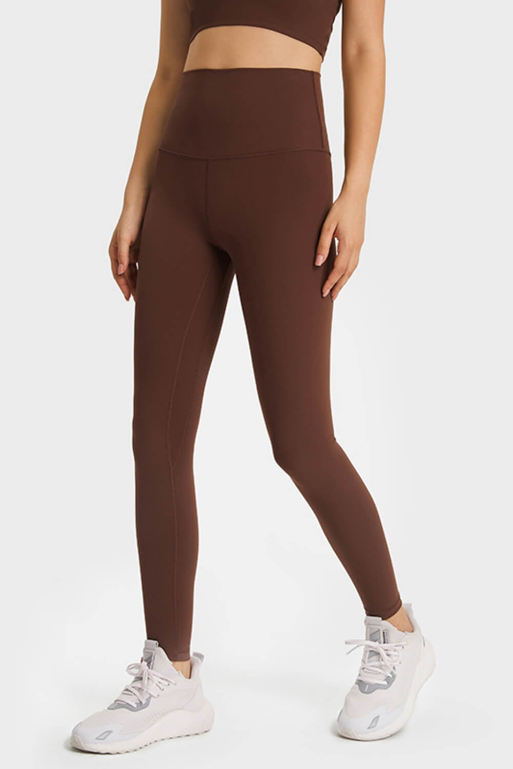 Model wears solid brown high-waist leggings with an elastic waistband, smooth fitted silhouette, long tapered legs, and visible seam construction.