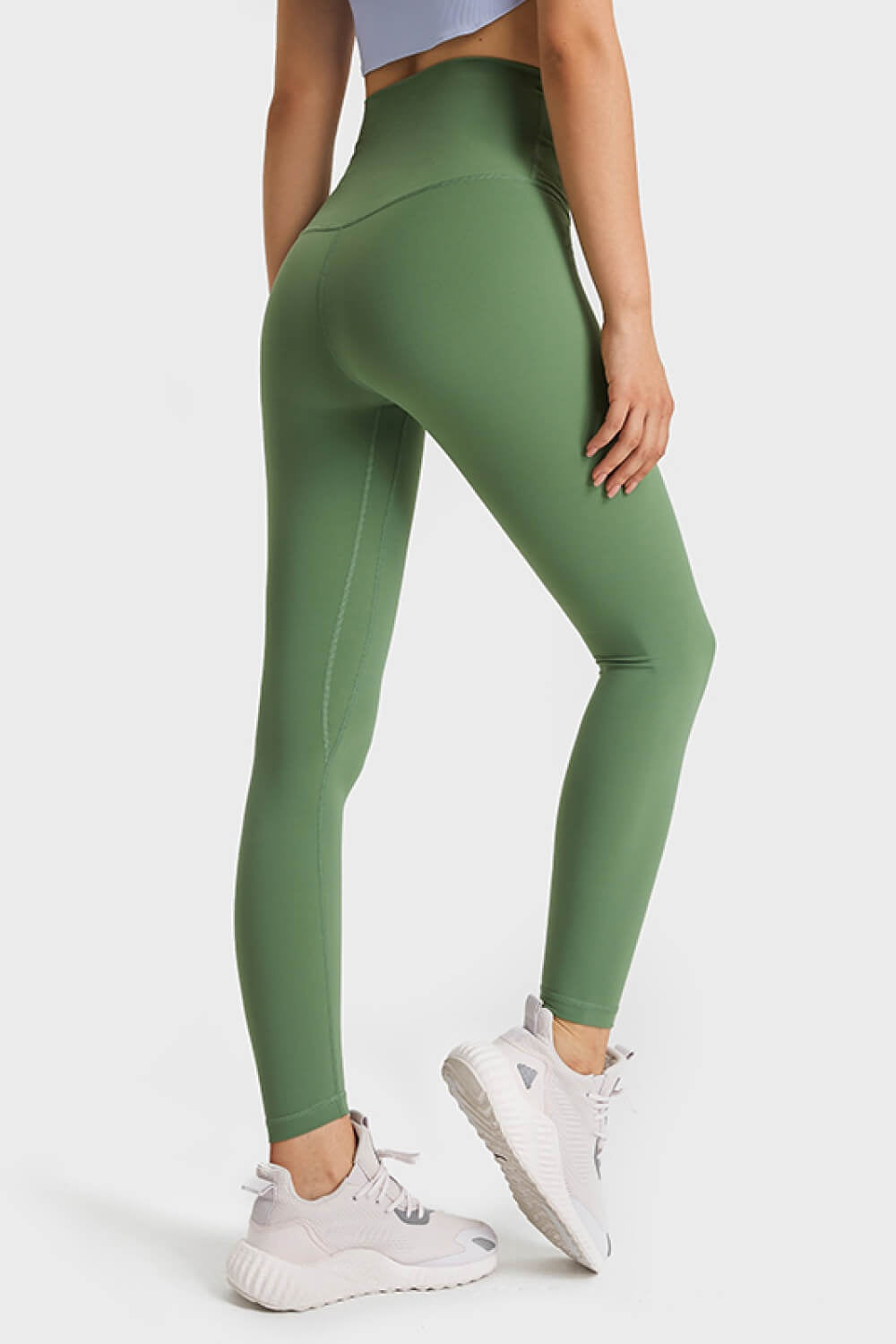 Model poses in green high-waist leggings with a smooth, sculpted silhouette, elastic waistband, and visible seam detailing, paired with white sneakers.