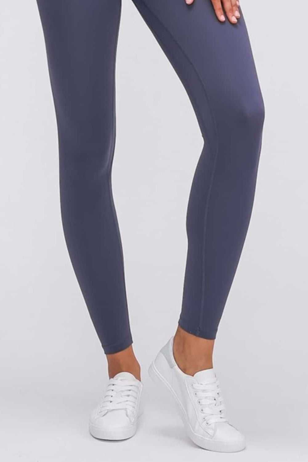 A model wears ultra soft solid navy leggings with a sleek, close-fitting ankle-length silhouette, smooth construction, and white sneakers while standing with one leg crossed.