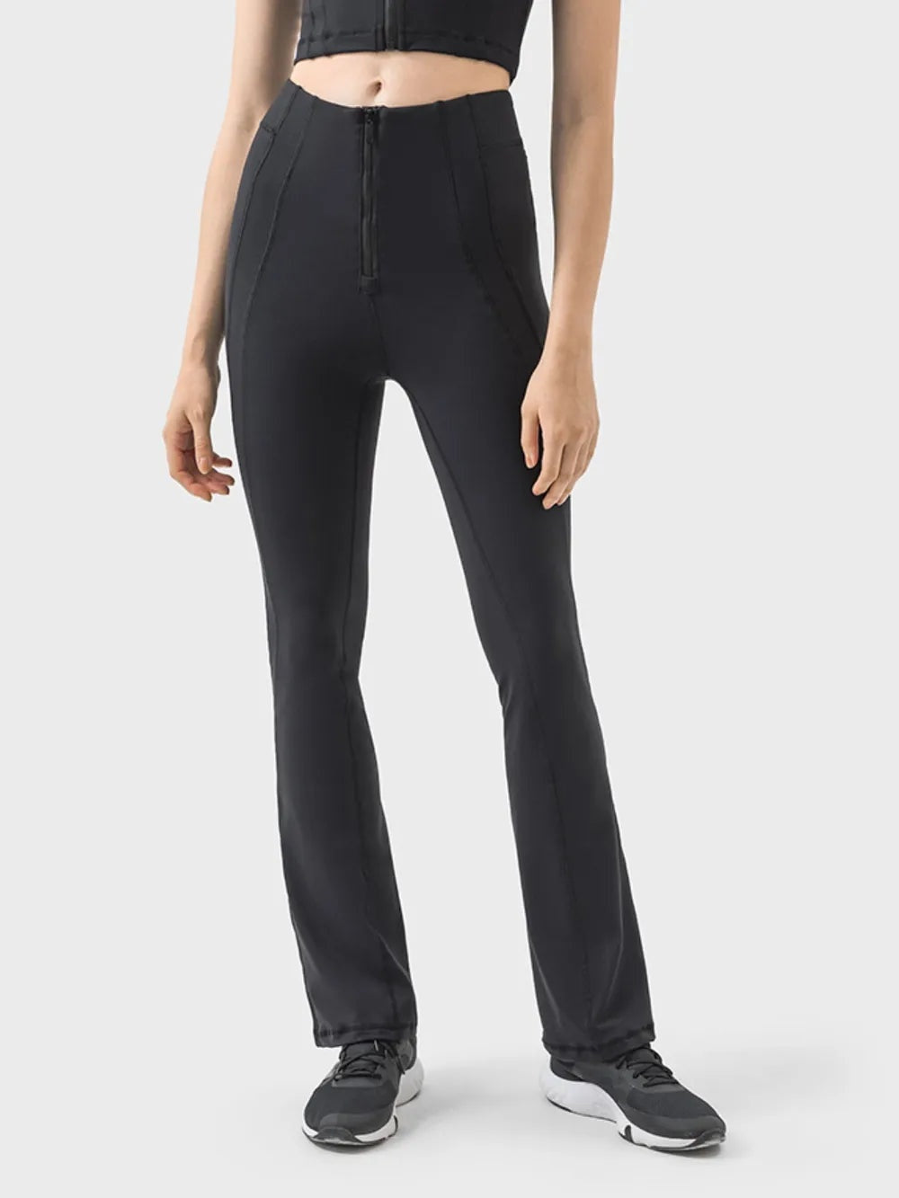 A model stands in black high-waist active pants with a front zipper, contoured seam detailing, and a sleek, fitted straight-leg silhouette.