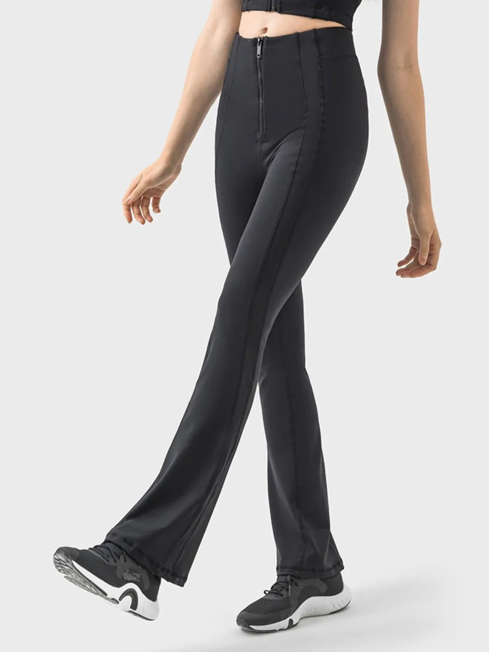 A model walks in black high-waist active pants with a front zipper, contoured seam panels, a fitted hip and thigh, and subtly flared legs.