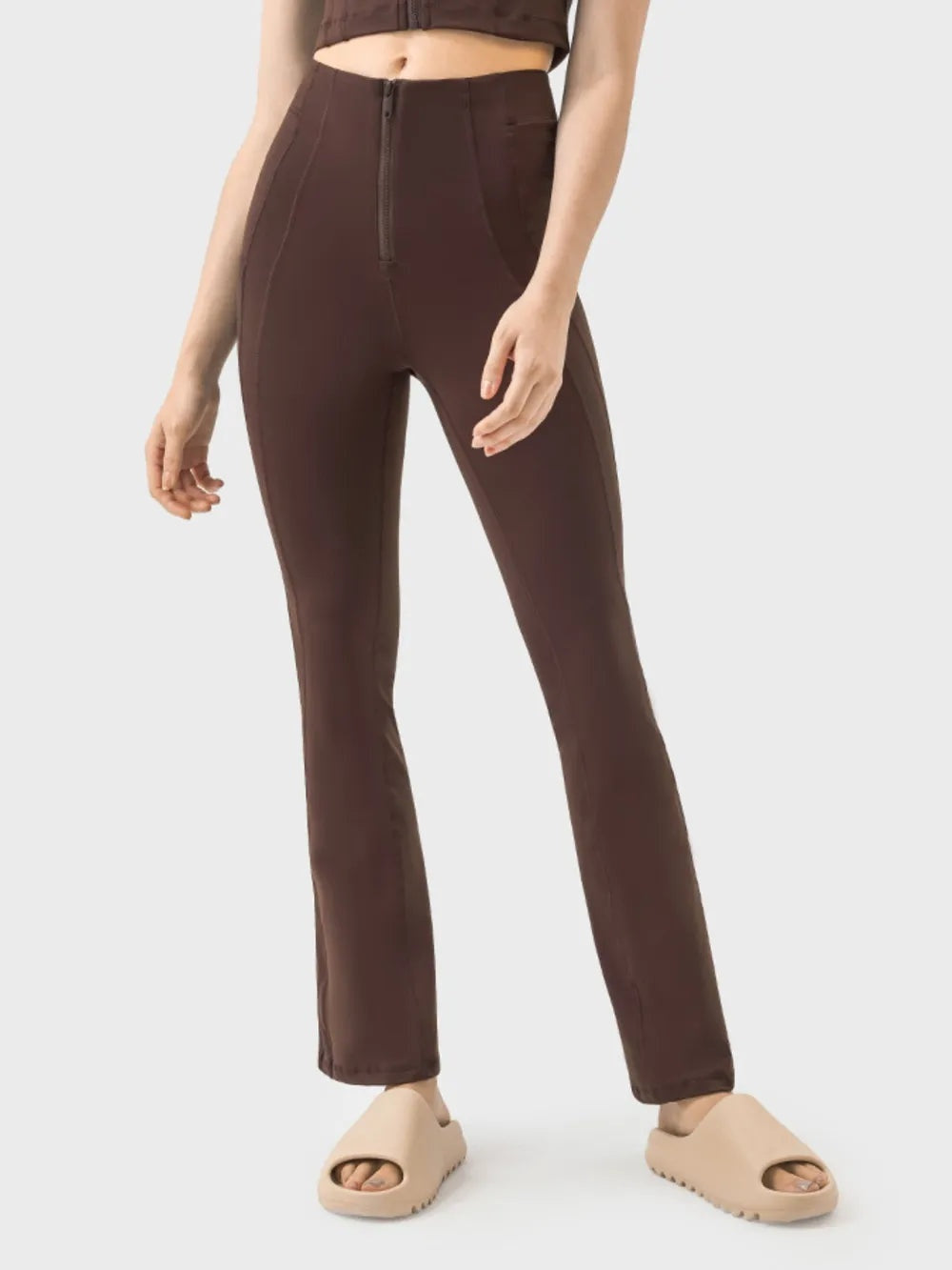 A model stands in dark brown high-waist active pants with a front zipper, slim fitted silhouette, subtle stretch, and straight legs with a slight flare.