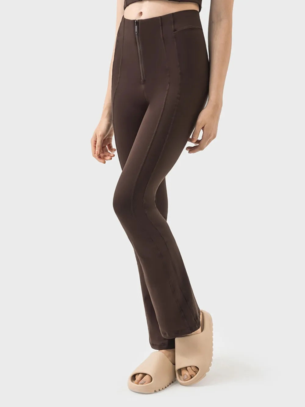 Model poses in dark brown high-waist active pants with a slim, slightly flared silhouette, front zipper detail, zippered pocket, and leg seam construction.