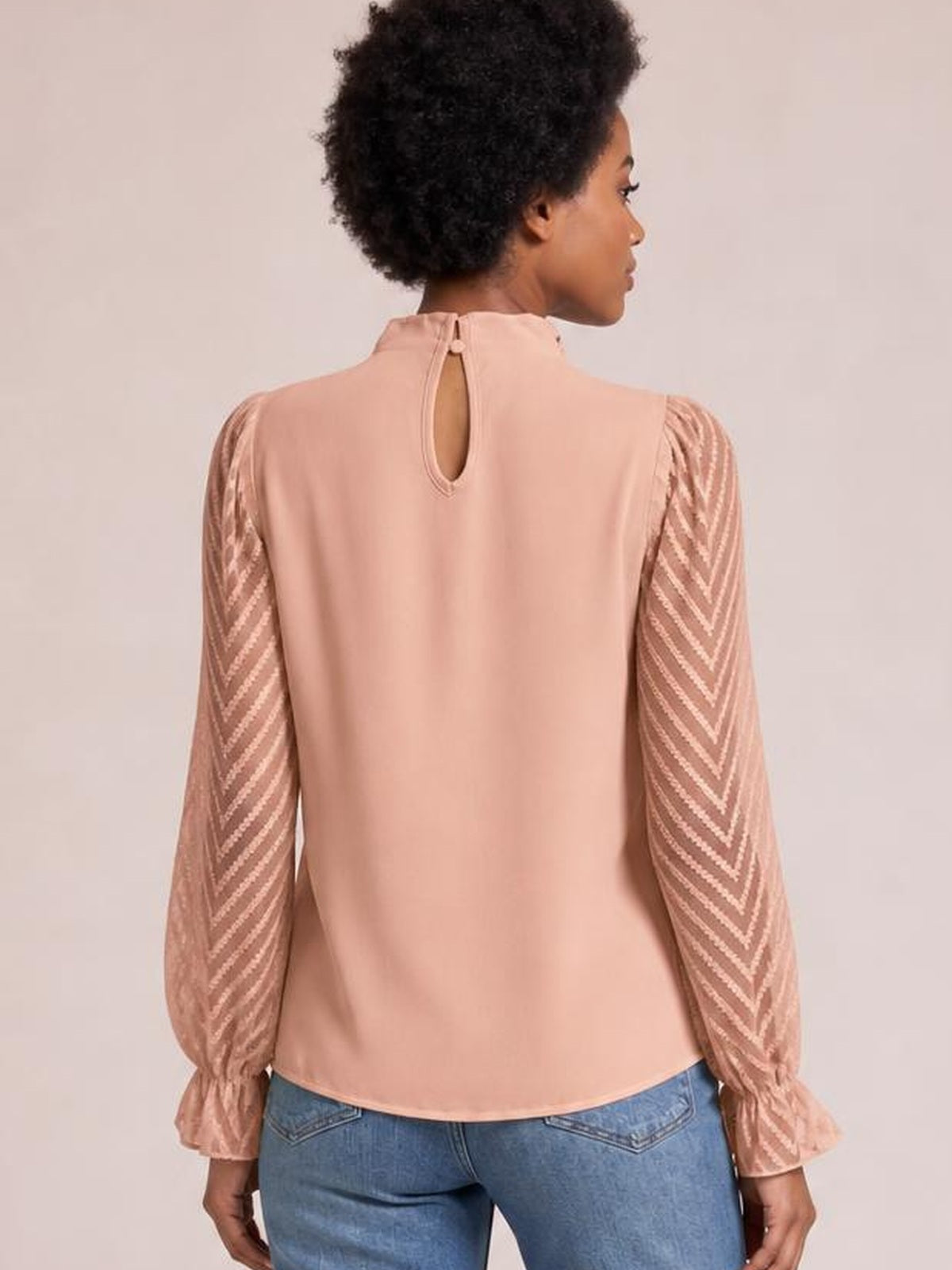 A model shows the back of a blush pink mock neck blouse with a keyhole button closure, sheer chevron sleeves, ruffled cuffs, and a relaxed silhouette.