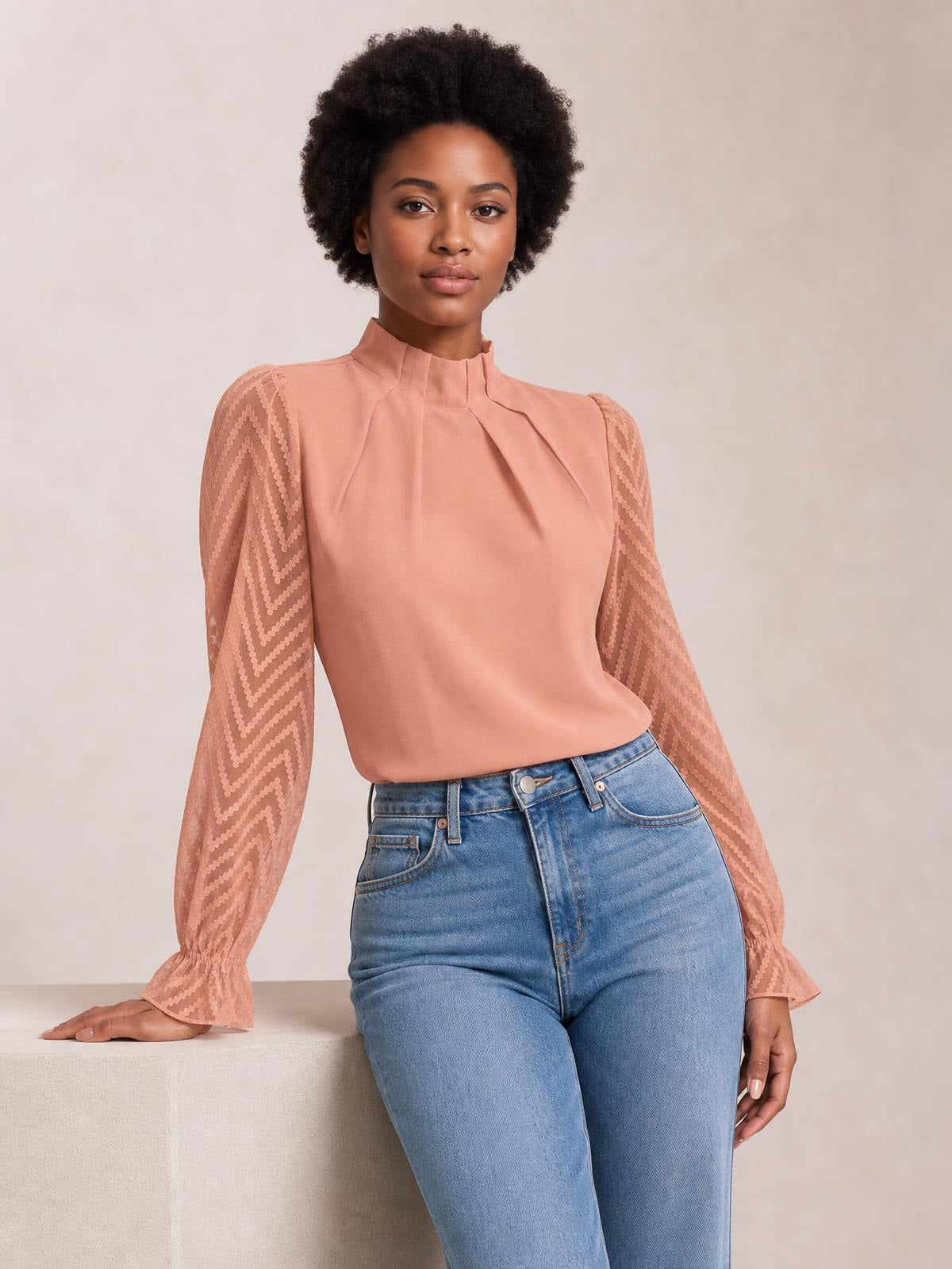 A model poses in a peach mock neck blouse with a smooth bodice, sheer patterned flounce sleeves, and gathered ruffled cuffs.
