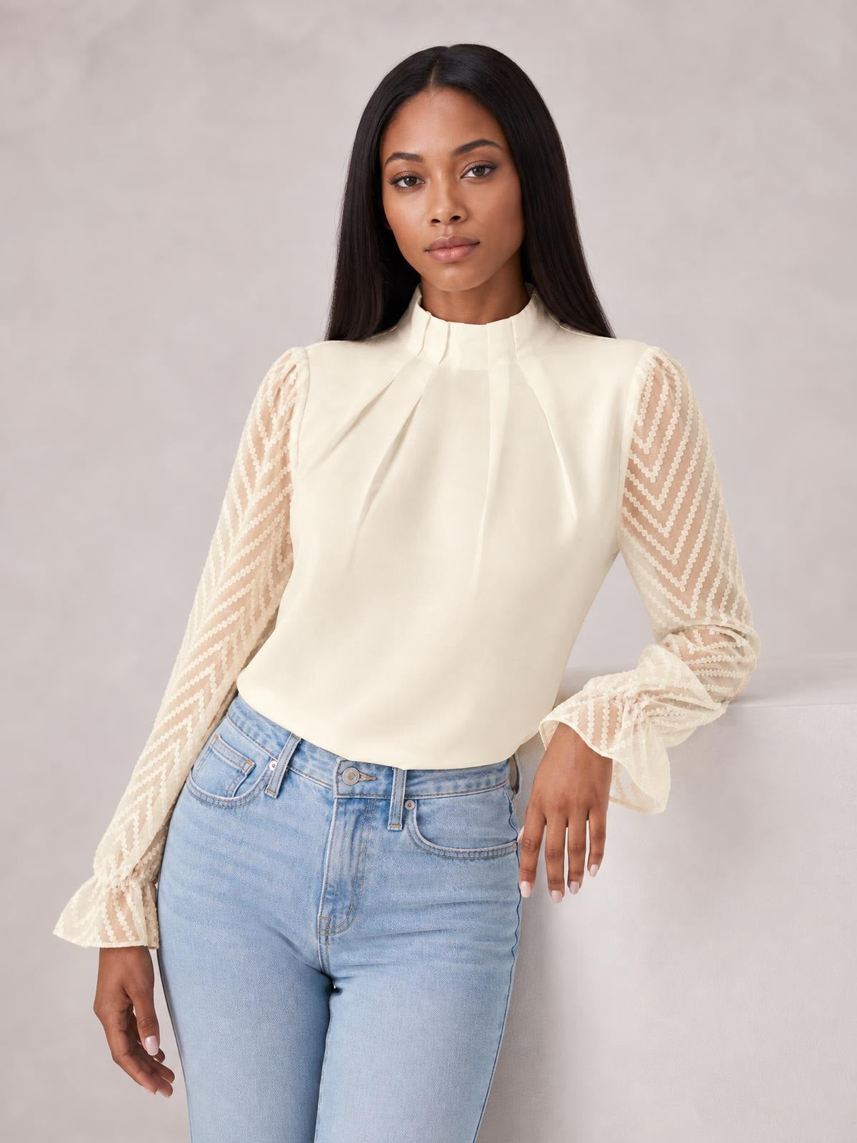 A model wears an ivory mock neck blouse with a smooth silhouette, sheer patterned sleeves, and ruffled flounce cuffs while standing with one hand at her hip.