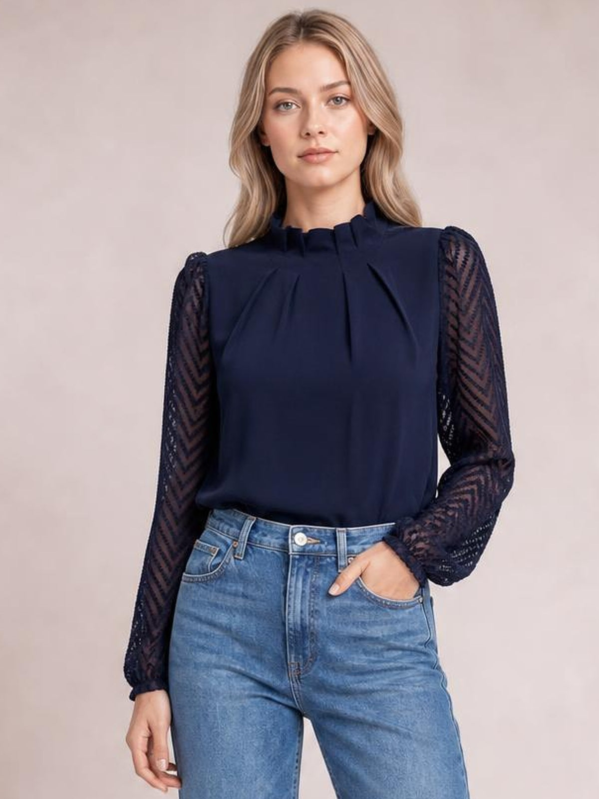 Model poses in a navy mock neck blouse with a ruffled collar, pleated front, sheer patterned flounce sleeves, and a relaxed silhouette tucked into jeans.