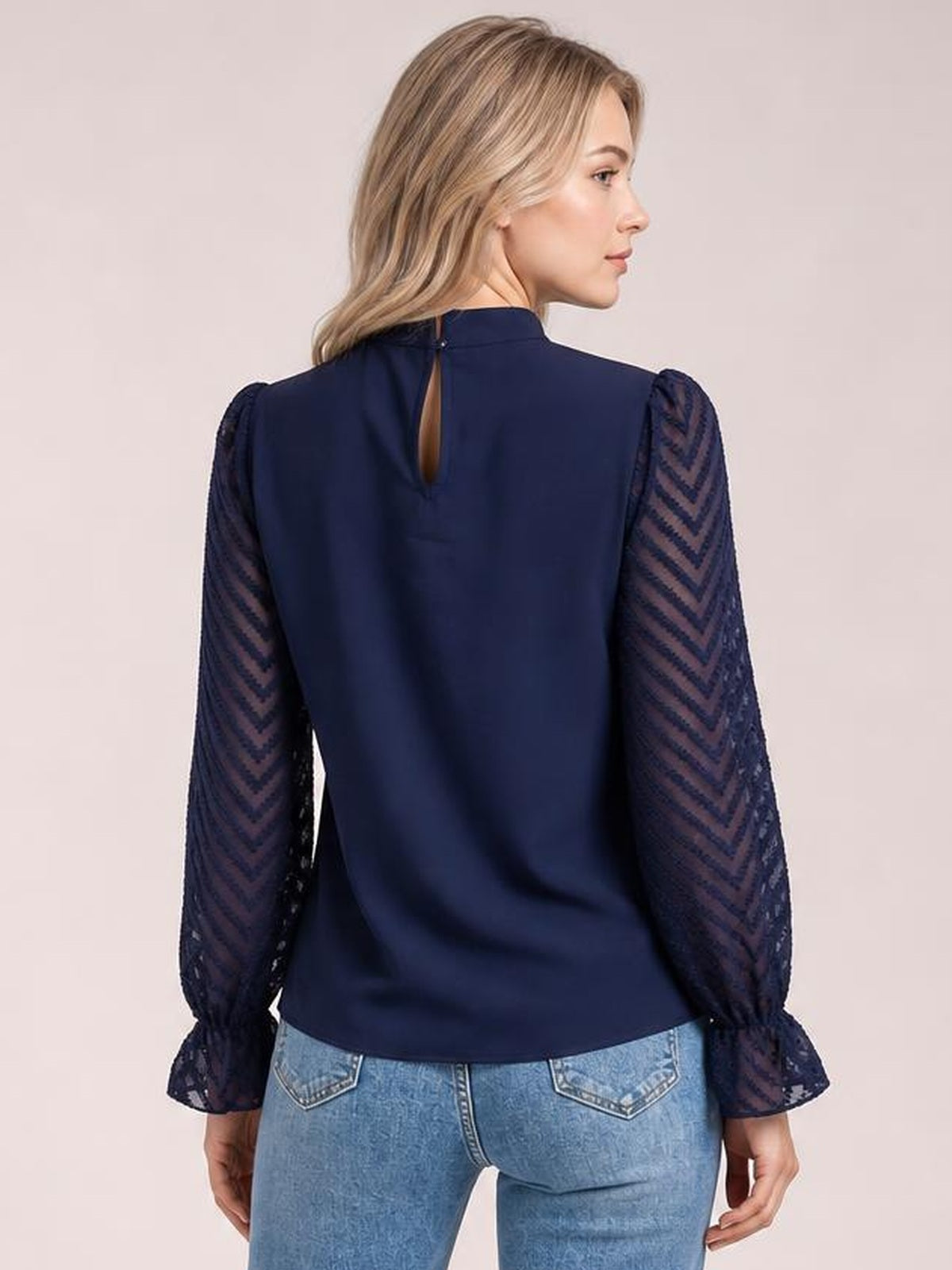 A model shows the back of a navy mock neck blouse with a keyhole closure, sheer patterned sleeves, flounce cuffs, and a relaxed straight silhouette.