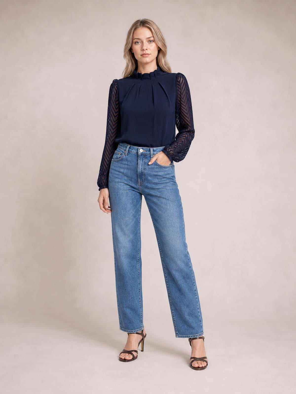 Model poses in a navy mock neck blouse with ruffled collar, pleated front, and sheer patterned flounce sleeves, styled with straight-leg jeans.