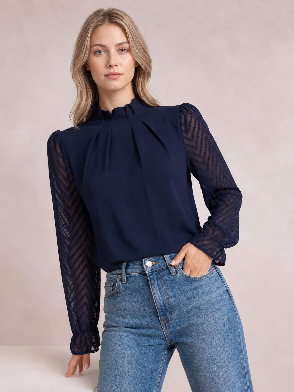 A model poses in a navy mock neck blouse with pleated front, sheer patterned long sleeves, gathered cuffs, and a relaxed silhouette tucked into jeans.