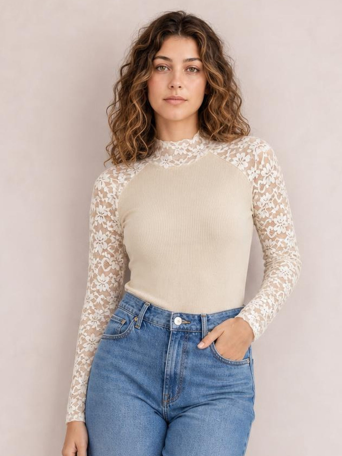 Model wears a beige mock neck long sleeve T-shirt with a fitted ribbed bodice and semi-sheer floral lace patchwork sleeves, one hand in her jeans pocket.