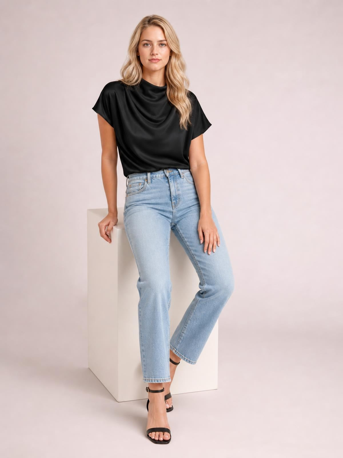 Model wears a black satin mock neck blouse with short sleeves, a softly draped neckline, relaxed silhouette, and tucked hem while leaning against a pedestal.
