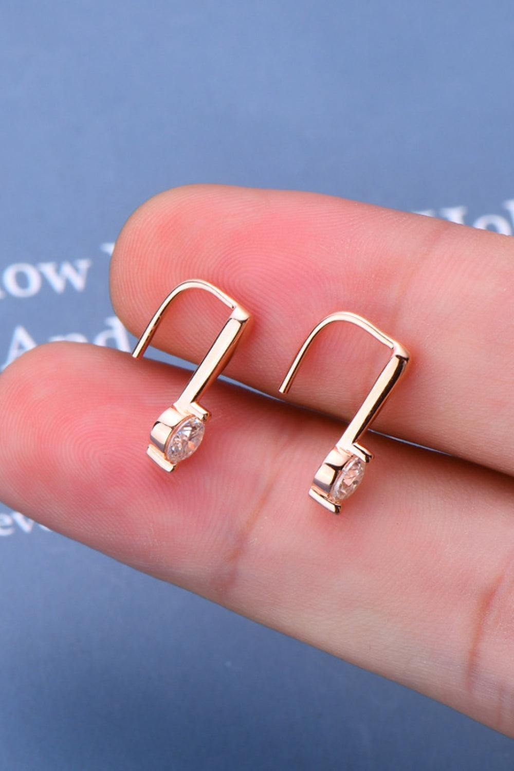 A pair of 925 sterling silver moissanite earrings with rose gold-plated rectangular drop settings and curved hook backs, shown in a hand.