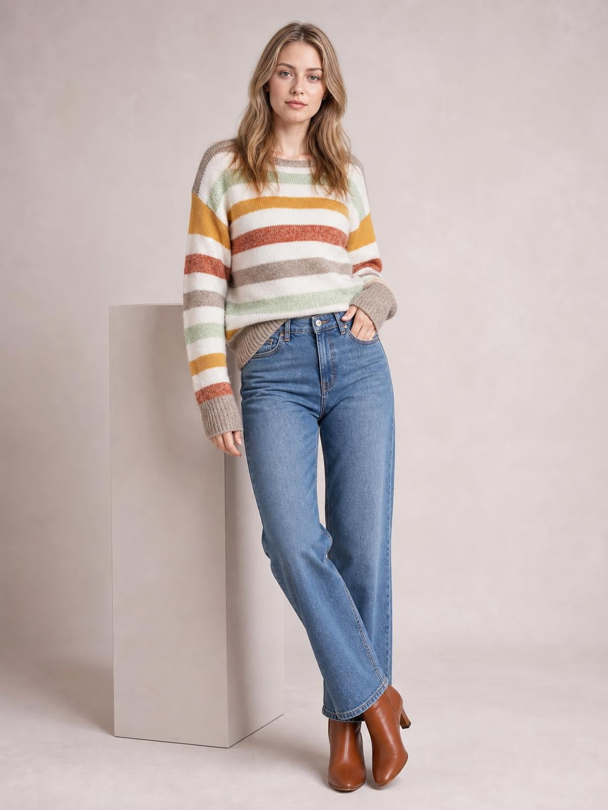 A model poses in a multicolour stripe crew neck sweater with drop shoulders, ribbed cuffs and hem, and a relaxed silhouette, one hand in her jeans pocket.