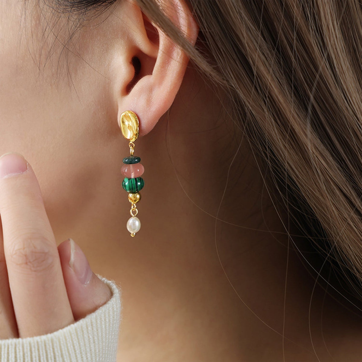 A model touches her ear, showcasing a gold-toned dangle earring with green and pink natural stone beads and a freshwater pearl drop, paired with a cream ribbed sleeve.