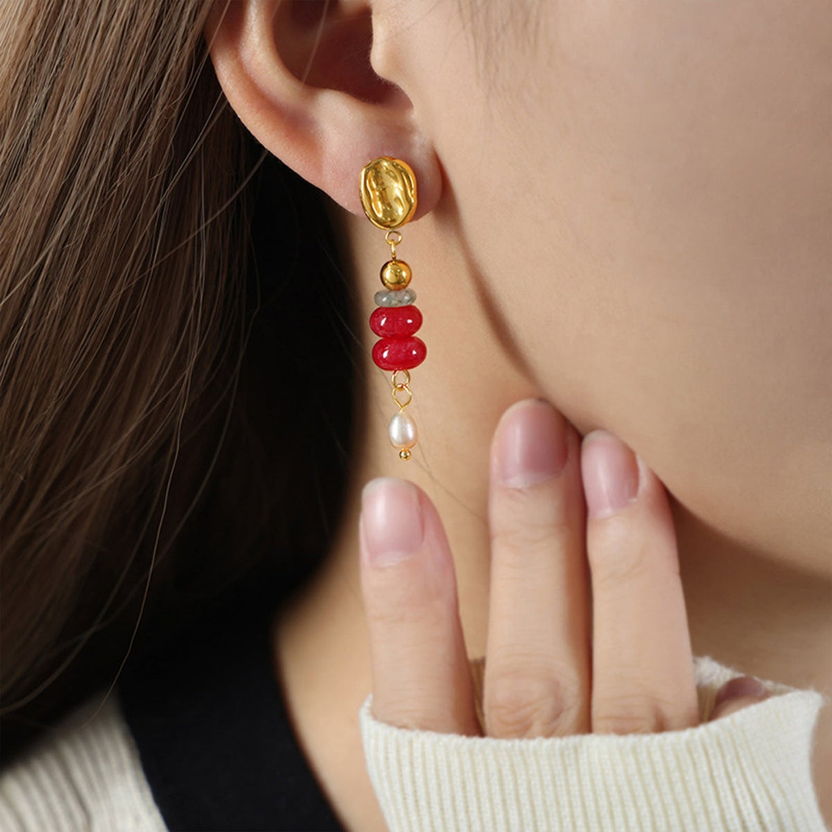 A close-up model touches her cheek while wearing a gold-plated natural stone freshwater pearl dangle earring with red beads, gray stone, and a cream ribbed sleeve.