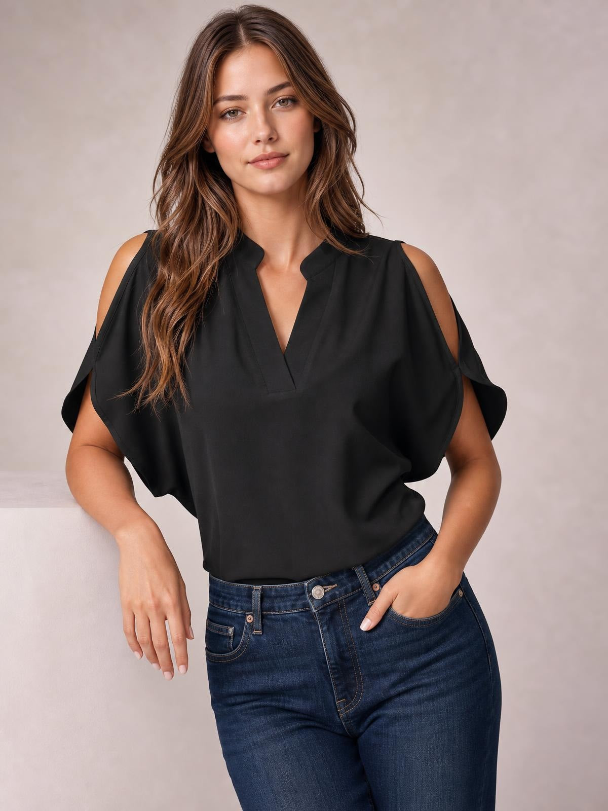A model poses with one hand in her jeans pocket wearing a black notched cold shoulder blouse with a relaxed, draped silhouette and flutter sleeves.