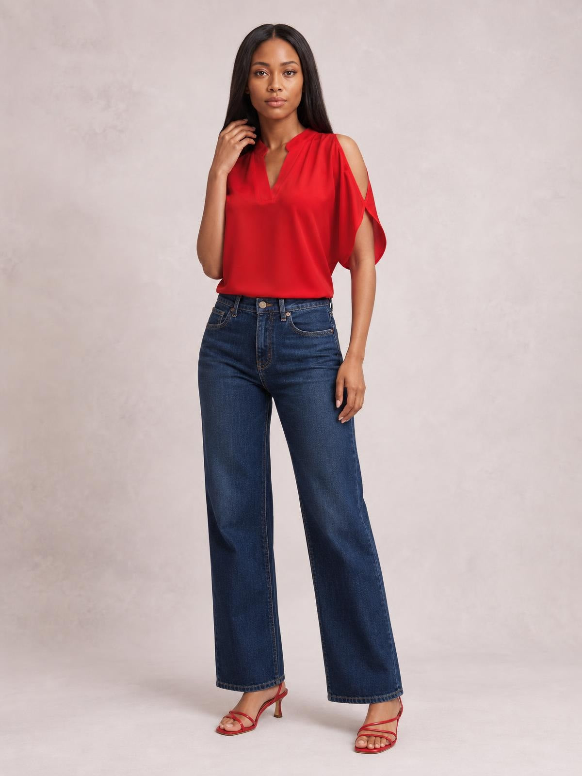 Model poses in a red notched cold shoulder blouse with a relaxed, fluid silhouette, V-neckline, and draped cutout sleeves over wide-leg jeans.