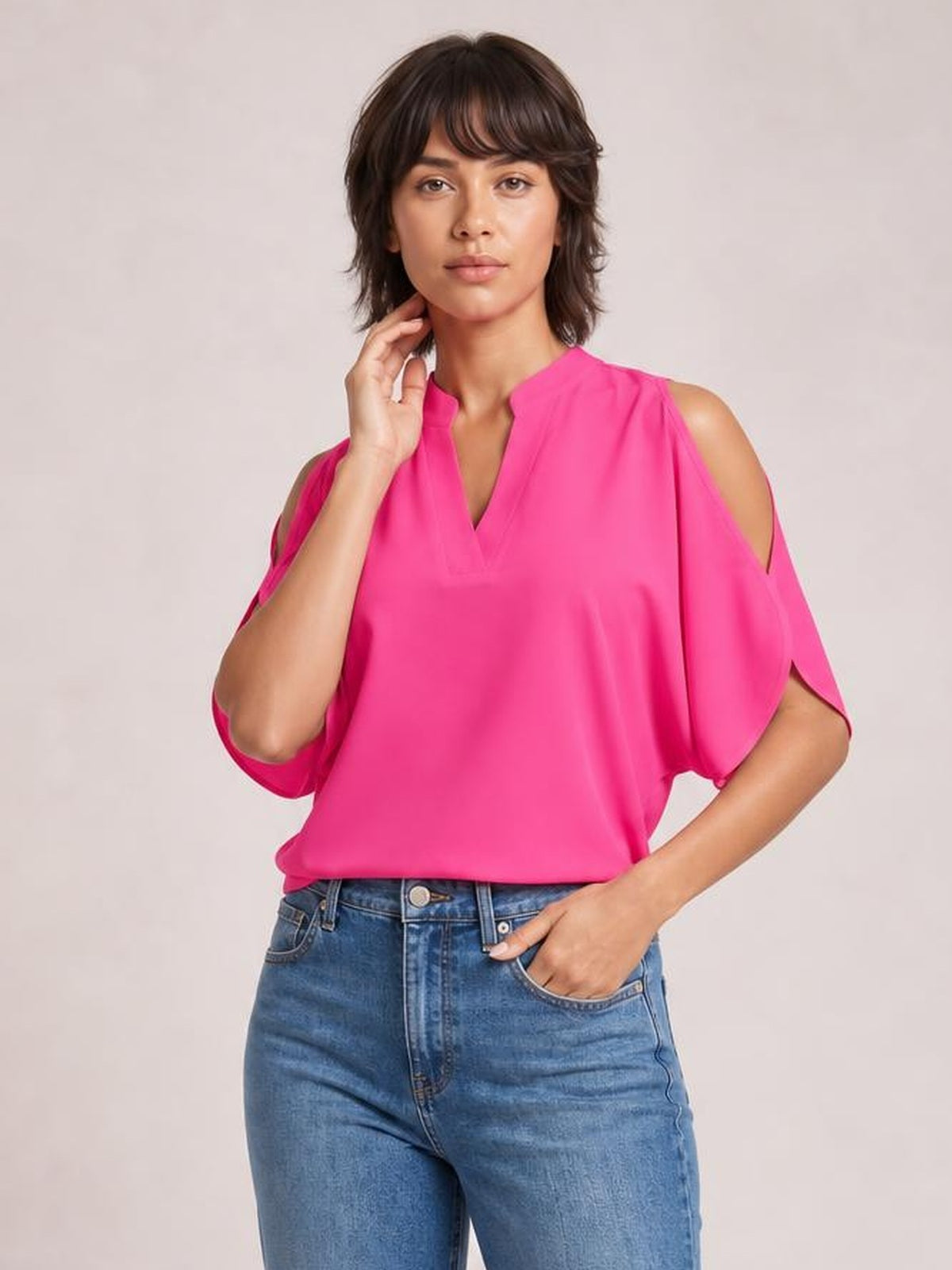 Model poses in a bright pink notched cold shoulder blouse with a relaxed silhouette, V-neckline, and flowing cutout sleeves.
