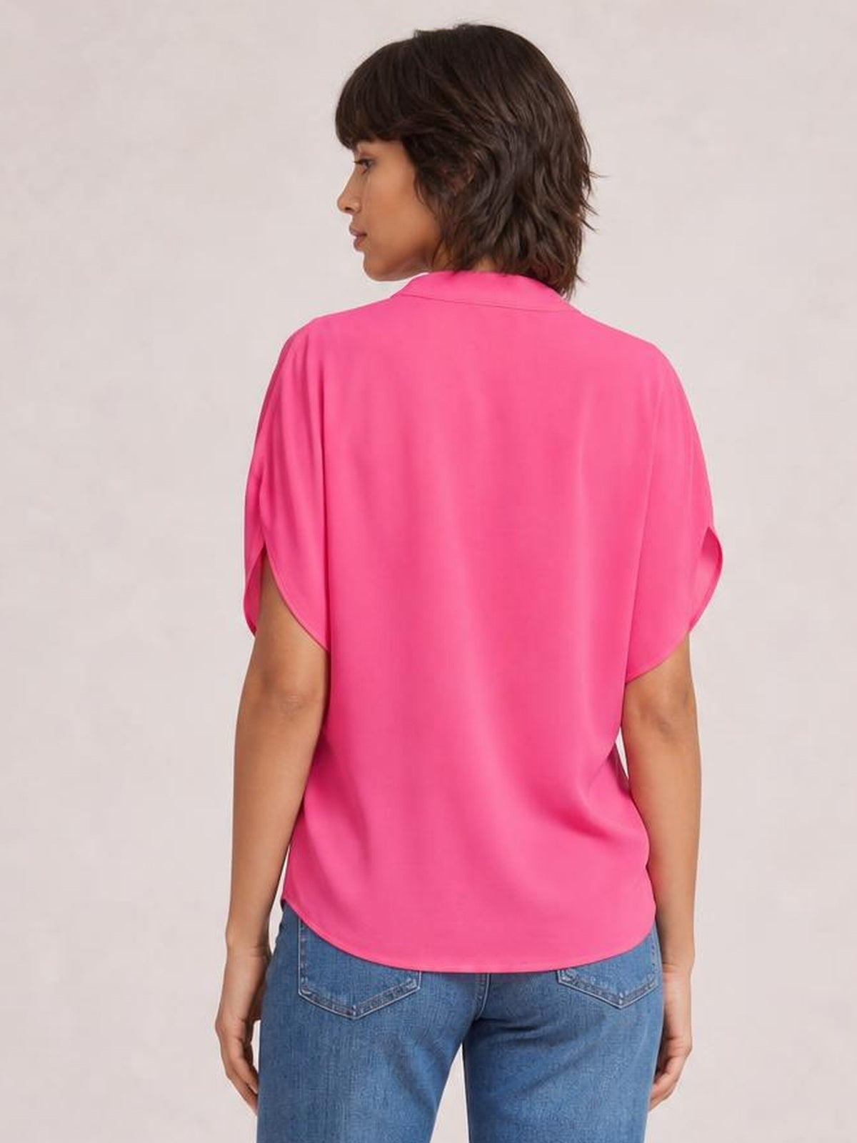 Model shown from behind in a bright pink notched cold shoulder blouse with a relaxed silhouette, collared neckline, slit sleeves, and curved hem.