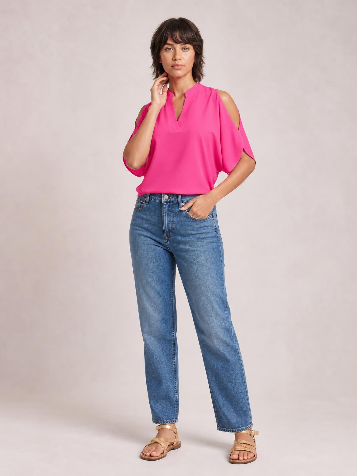 Model wearing a bright pink notched cold shoulder blouse with a relaxed silhouette, V-neckline, loose short sleeves, and cutout shoulders, posing with one hand at her neck.