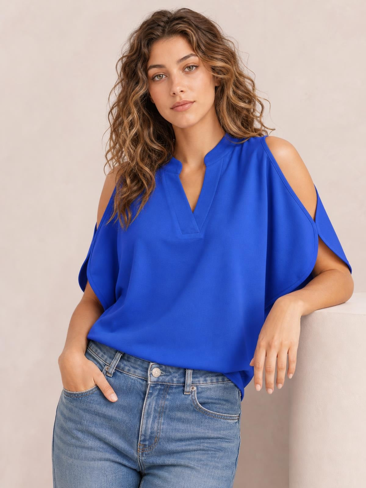 Model wears a bright blue notched cold shoulder blouse with a relaxed silhouette, short sleeves, and a loose drape, posing with one hand in her jeans pocket.