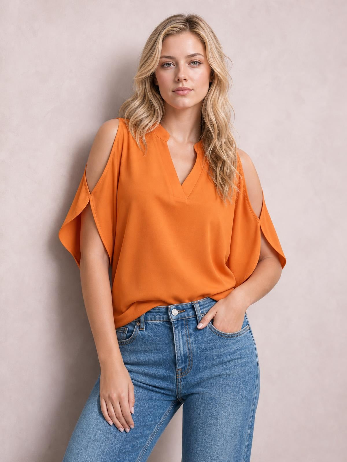 Model poses in an orange notched cold shoulder blouse with a relaxed silhouette, V-neckline, and flowing cutout sleeves tucked into blue jeans.