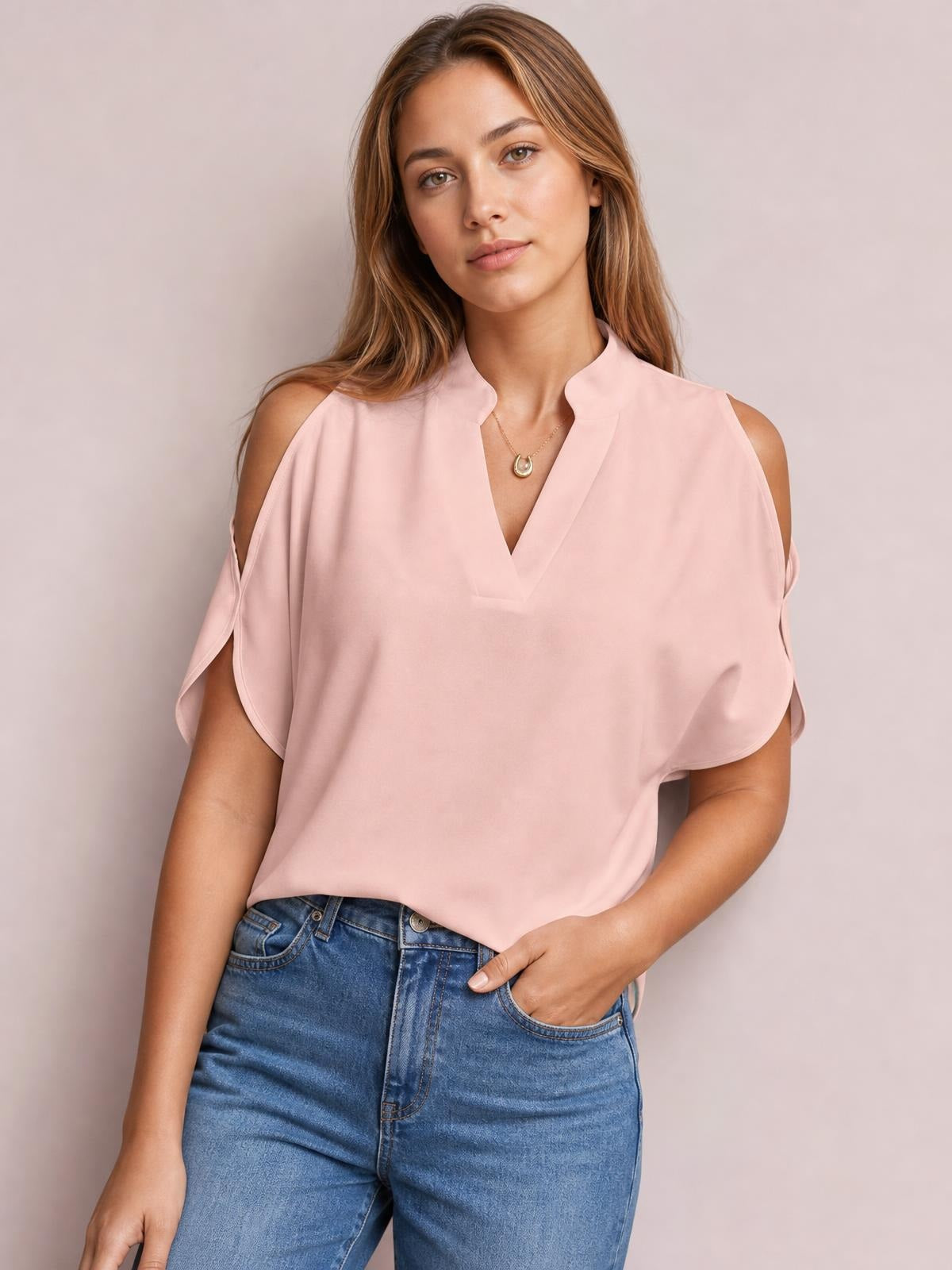 Model poses in a pink notched cold shoulder blouse with a relaxed silhouette, V-neckline, and draped short sleeves, paired with jeans.
