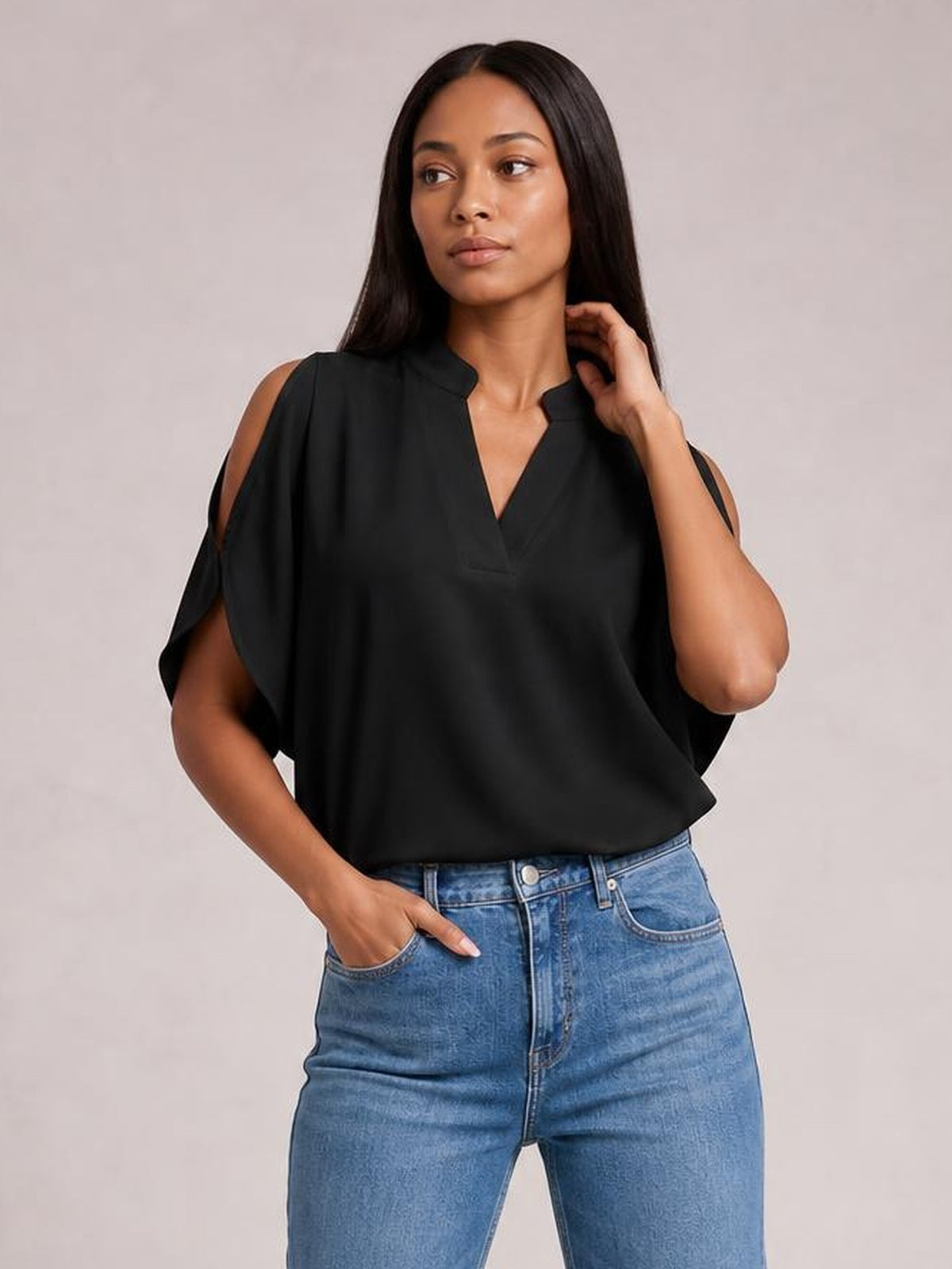 Model wearing a black notched cold shoulder blouse with relaxed half sleeves and a loose silhouette, posing with one hand in her jeans pocket and the other near her hair.