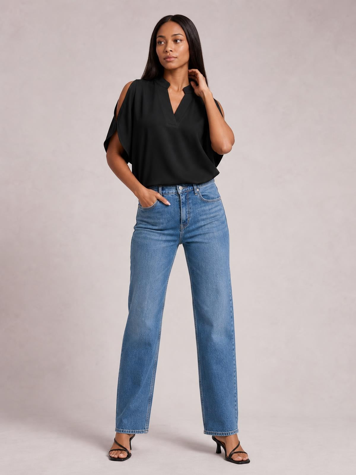 Model wearing a black notched cold shoulder blouse with half sleeves, a relaxed silhouette, and a loose drape, posing with one hand in her jeans pocket.