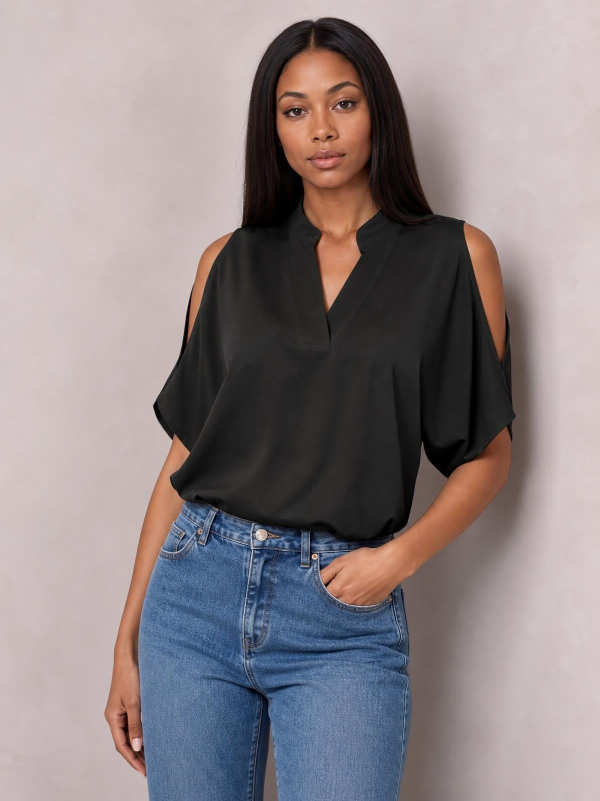 Model wears a black notched cold shoulder blouse with half sleeves, relaxed silhouette, and flowing opaque construction, posing with one hand in her jeans pocket.