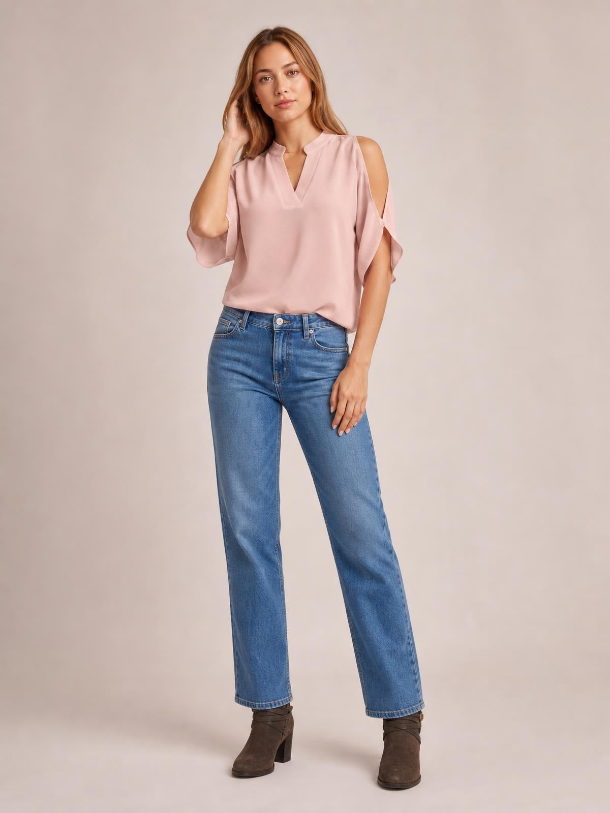 Model poses in a blush pink notched cold shoulder blouse with flowing half sleeves and a relaxed silhouette, paired with straight-leg blue jeans.