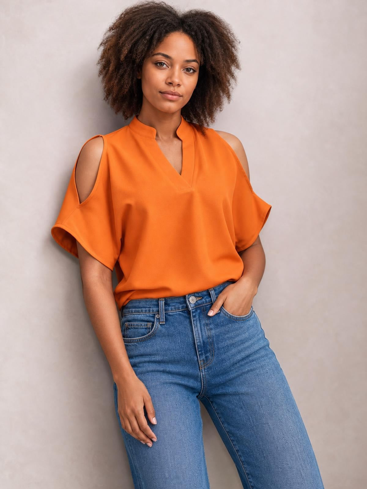 A model wears an orange notched cold shoulder blouse with short half sleeves, shoulder cutouts, a relaxed silhouette, and a V-shaped neckline.