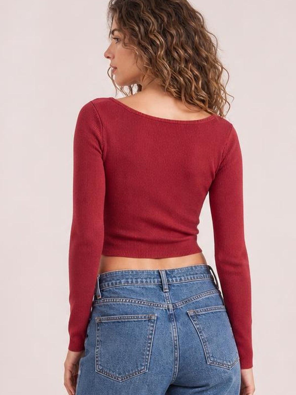 A model poses turned away in a fitted deep red long sleeve T-shirt with a wide neckline, fine ribbed texture, cropped hem, and slim silhouette.
