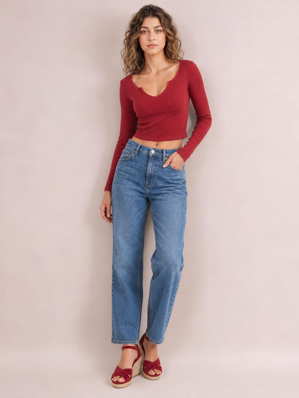 A model poses in a fitted burgundy notched long sleeve T-shirt with a ribbed texture, scoop neckline, and cropped silhouette, one hand in her jeans pocket.