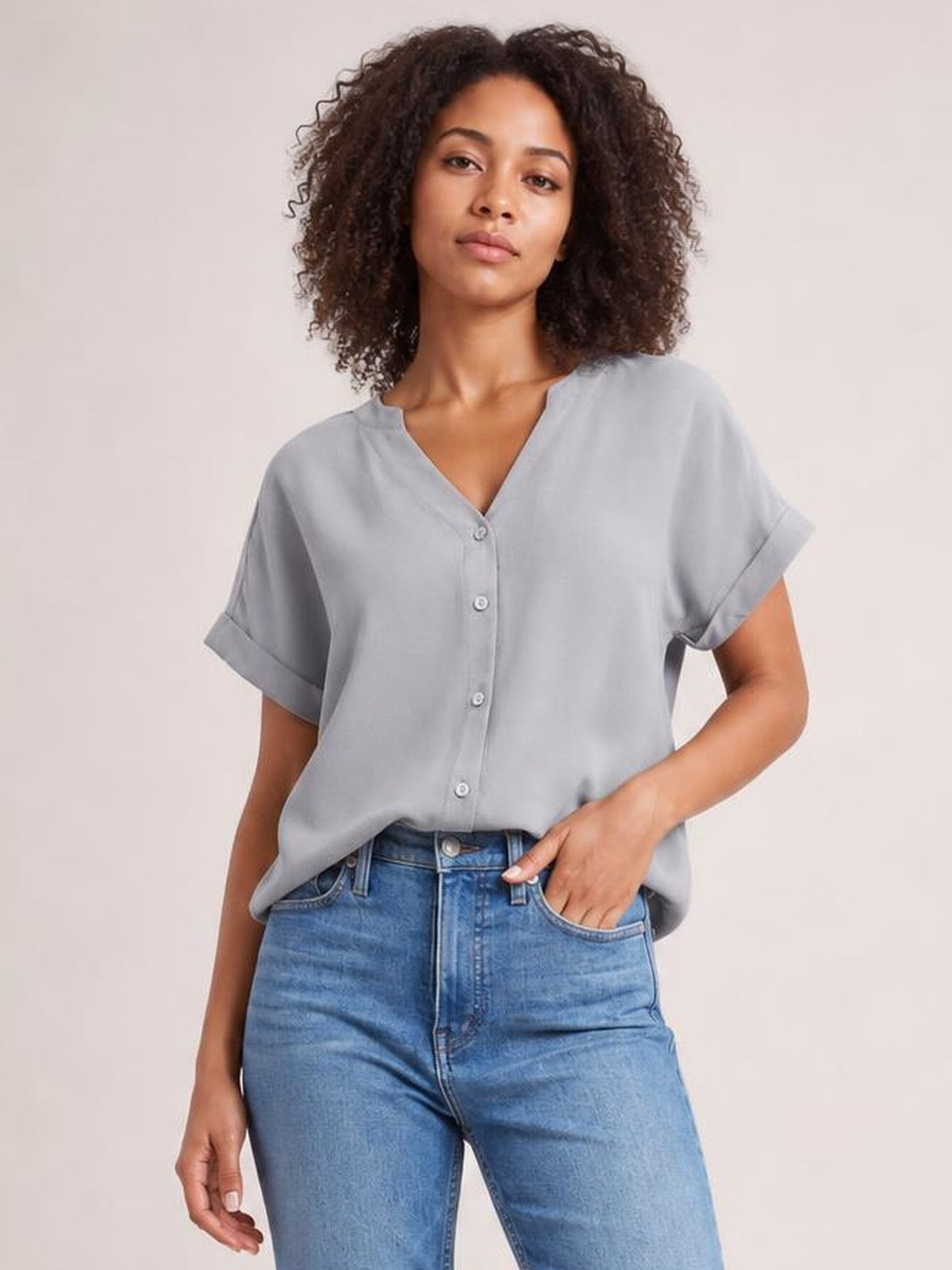 Model wearing a solid light gray notched neck shirt with button front, short cuffed sleeves, and a relaxed regular-length silhouette, posing with one hand in her jeans pocket.