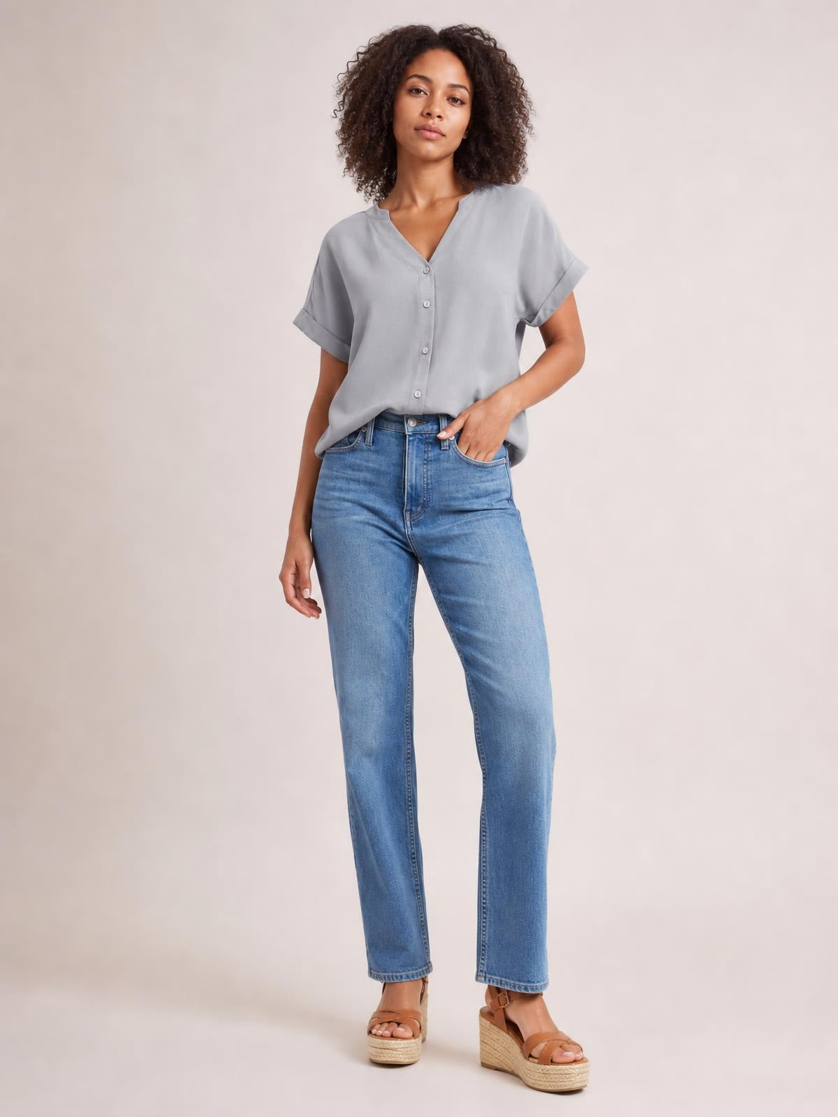 Model wearing a solid light gray notched neck shirt with button front, rolled cuffed short sleeves, and a relaxed regular-length silhouette, one hand in her jeans pocket.