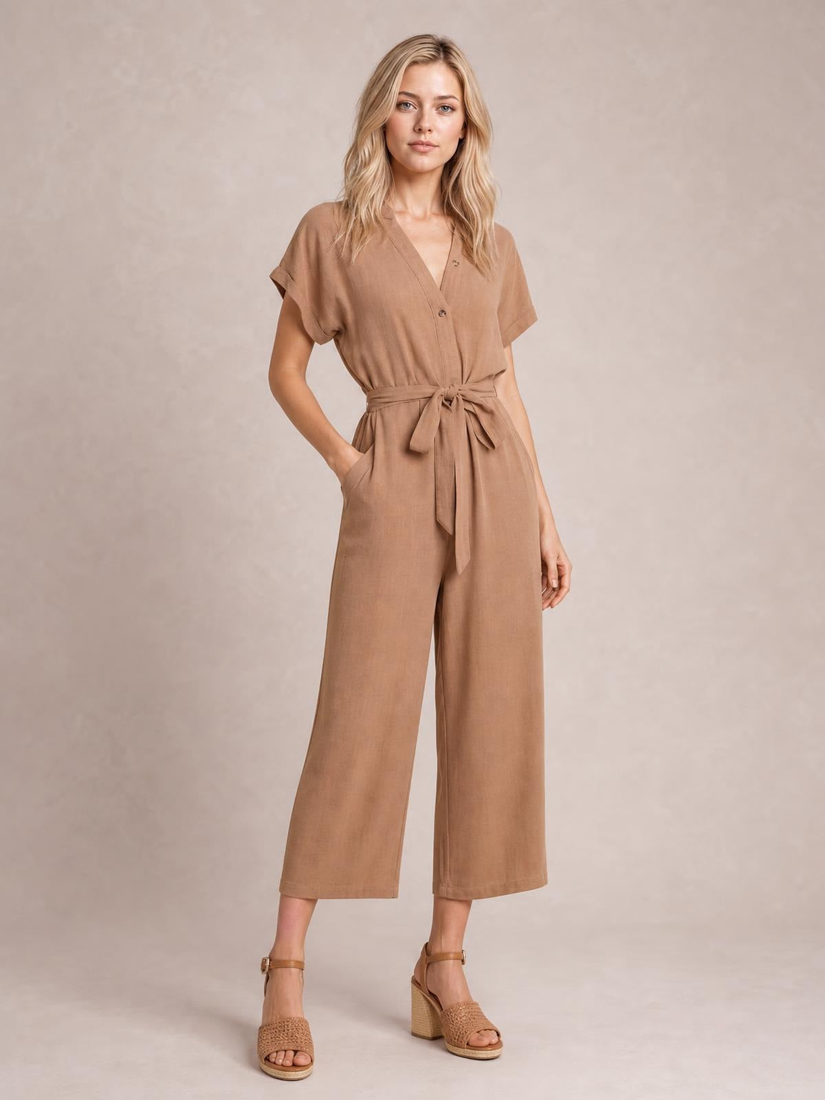 Model wearing a camel notched short sleeve tie waist jumpsuit with front buttons, pockets, and wide cropped legs.