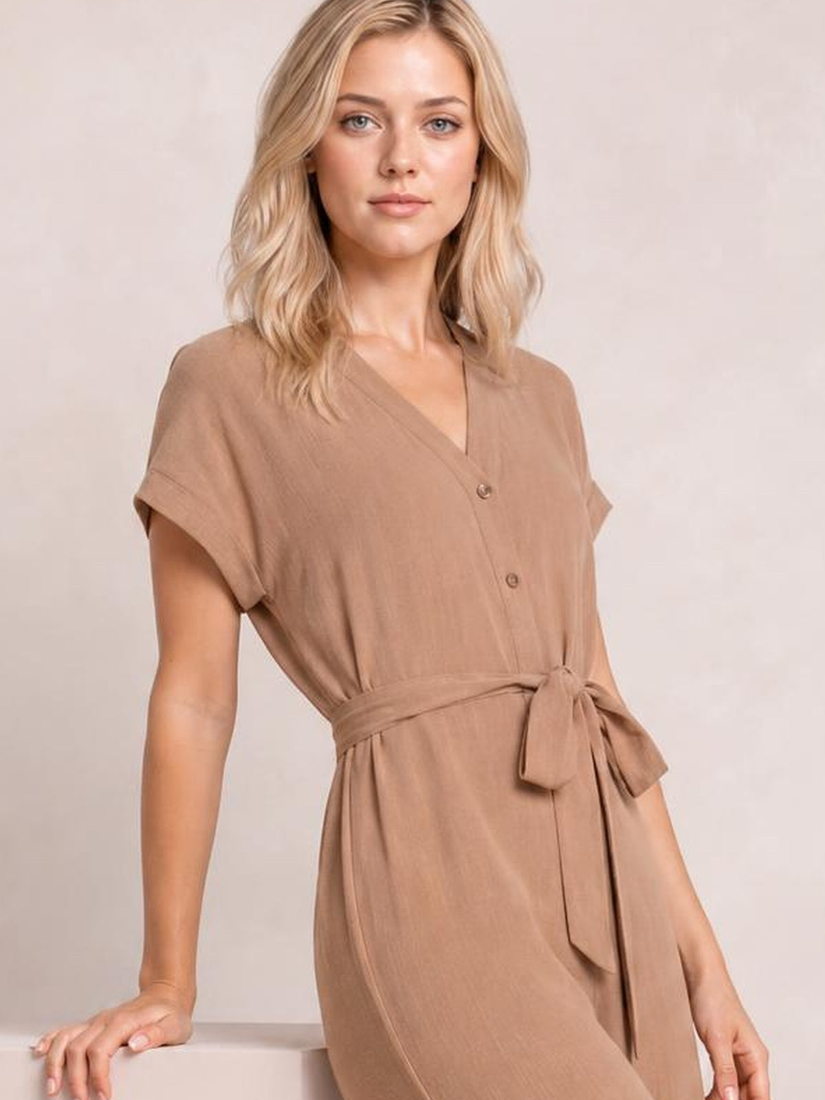 Model leans with one hand on a surface wearing a taupe notched short sleeve jumpsuit with front buttons, a tied waist, and a relaxed silhouette.