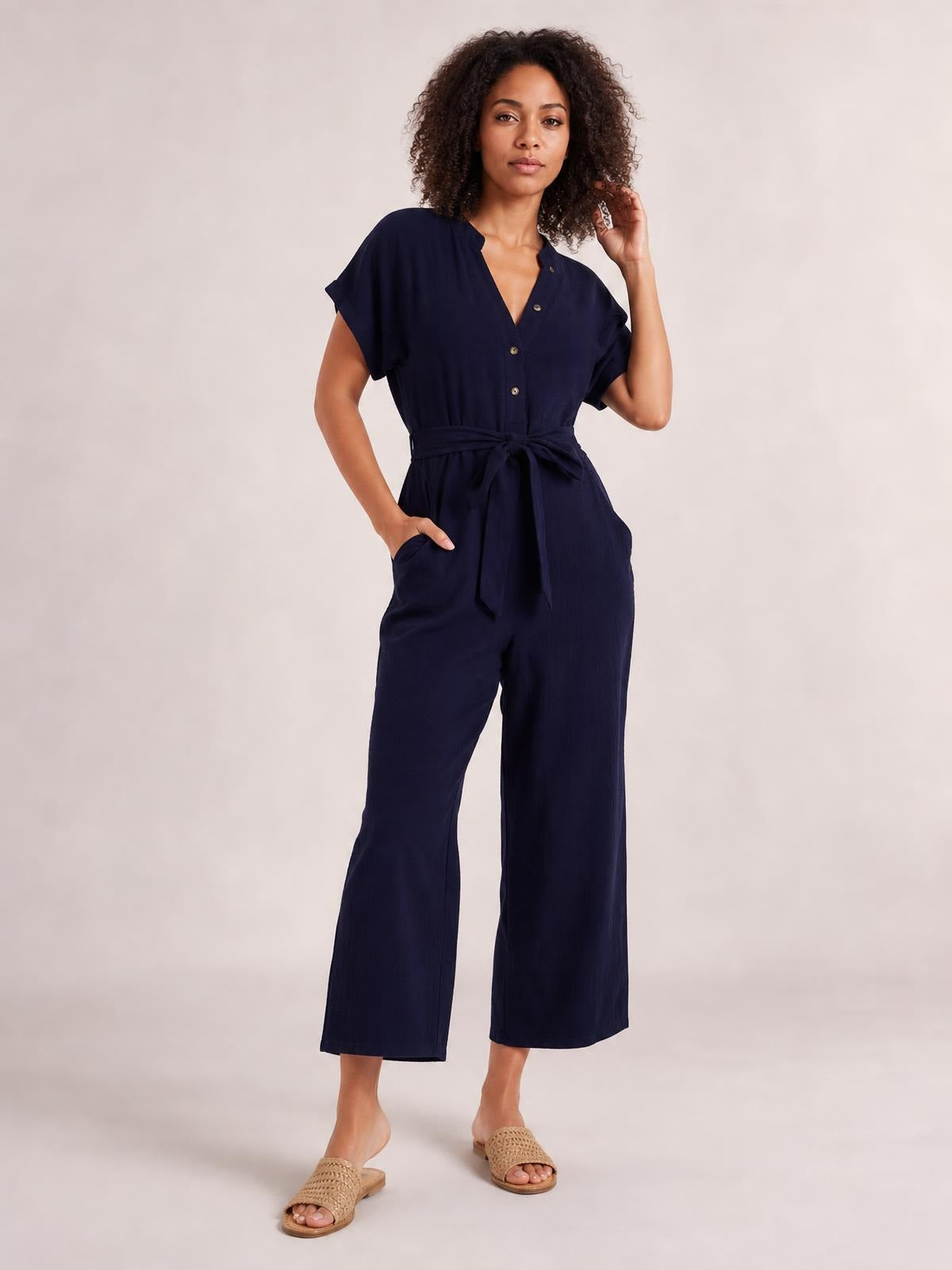 A model poses in a navy notched short sleeve jumpsuit with button front, tied waist, side pockets, and relaxed wide-leg trousers.
