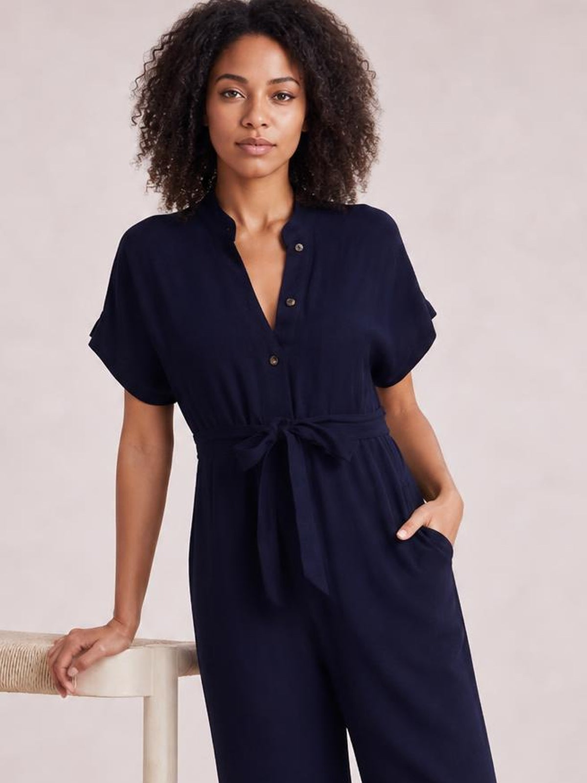 Model wearing a navy notched short sleeve tie waist jumpsuit with front buttons, wide-leg silhouette, and a visible side pocket.
