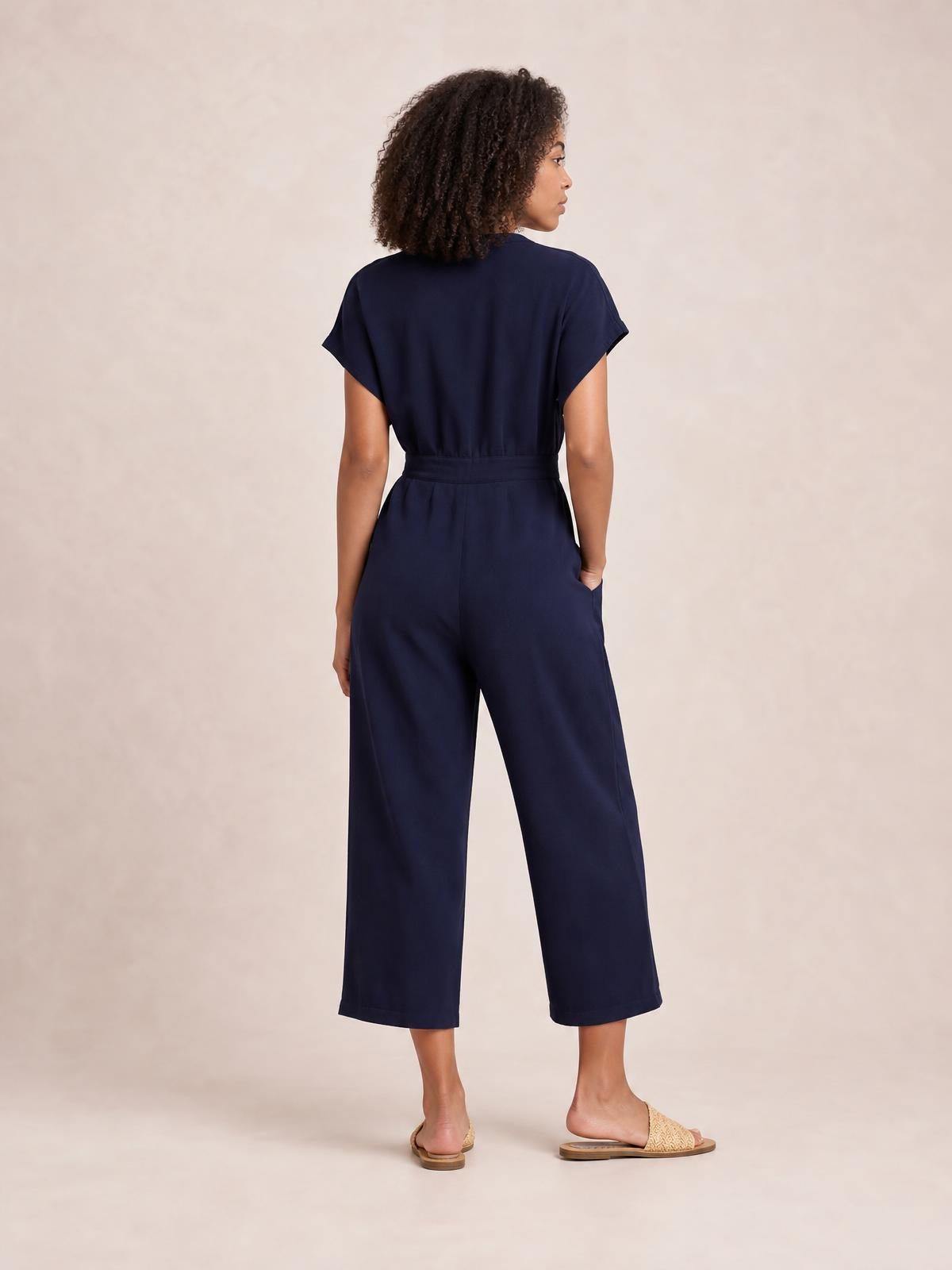 Model shown from behind in a navy notched short sleeve jumpsuit with a defined tie waist, cropped wide-leg silhouette, and side pockets.