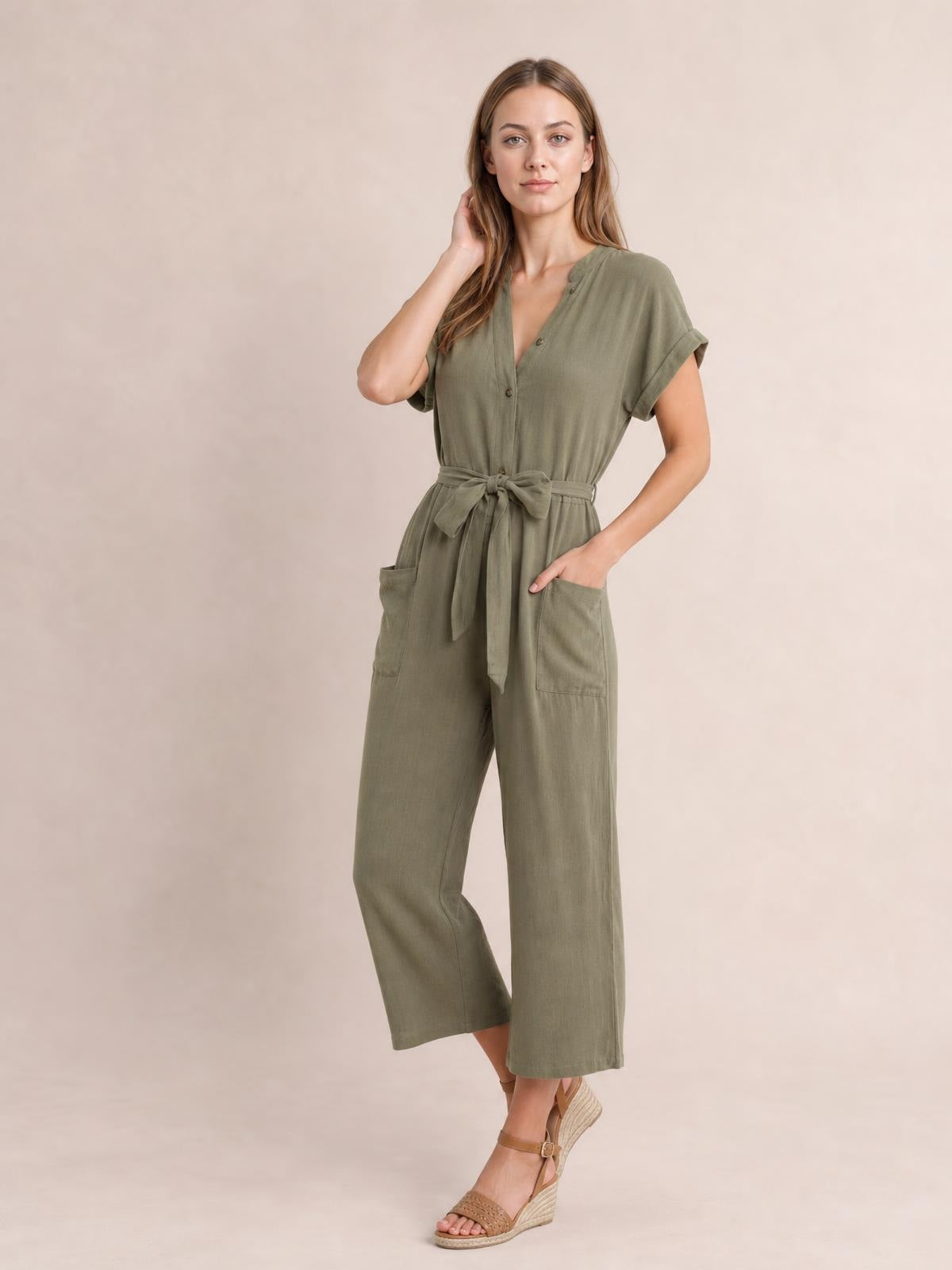 Model wearing an olive notched short sleeve tie waist jumpsuit with button front, wide cropped legs, and practical patch pockets, posing with one hand in a pocket.