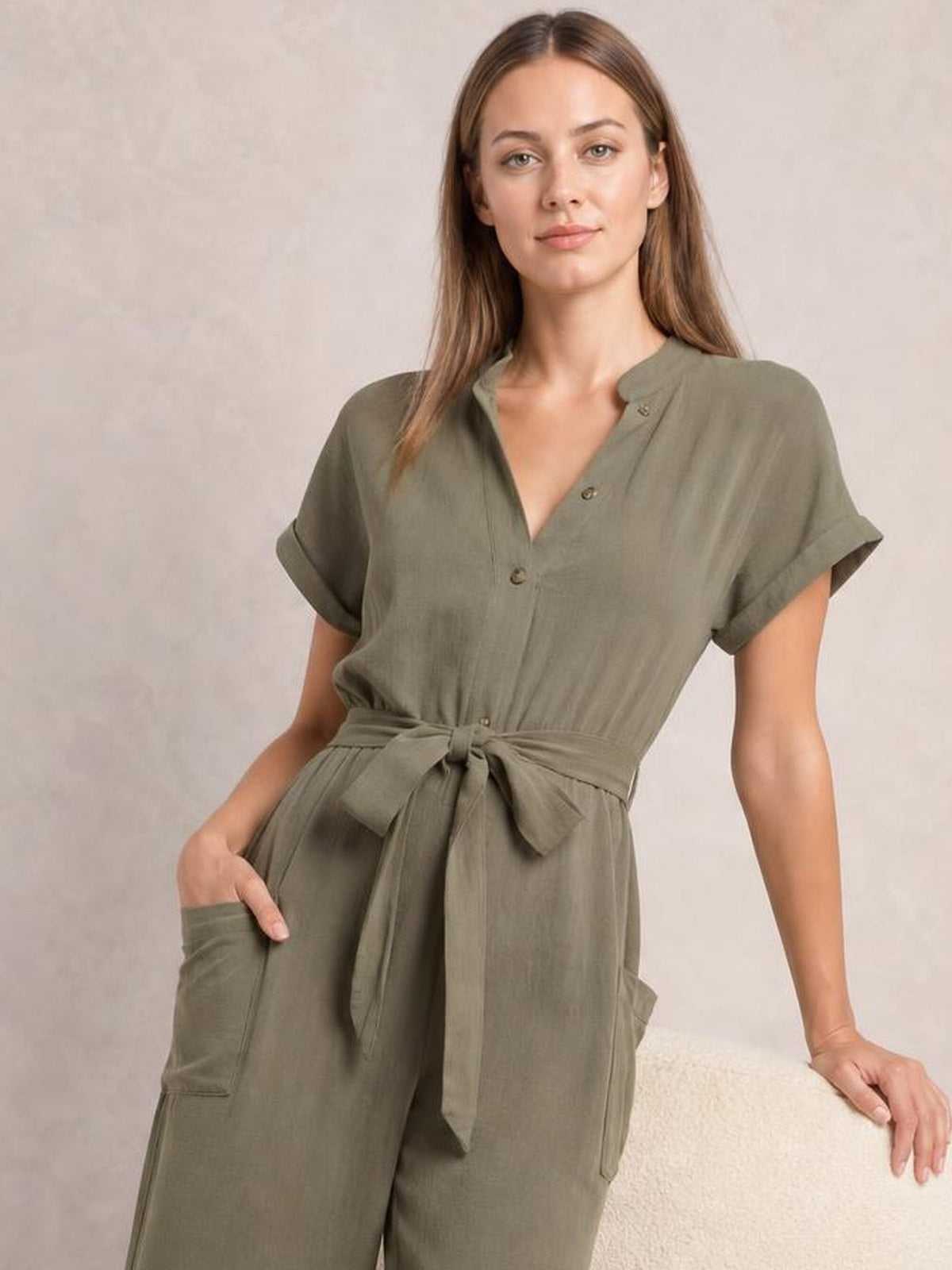 A model wears an olive green notched short sleeve jumpsuit with button front, tie waist, wide-leg silhouette, and utility pockets while leaning with one hand tucked in.